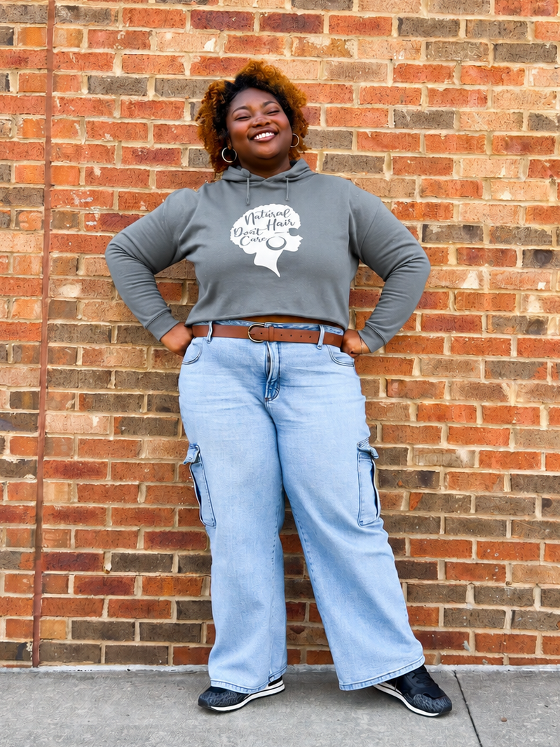 A woman stands against a red brick wall wearing a gray graphic hoodie with a white woman with an afro design and the text 'Natural Hair Don't Care', light blue wide-leg cargo jeans, a brown belt, and black sneakers.