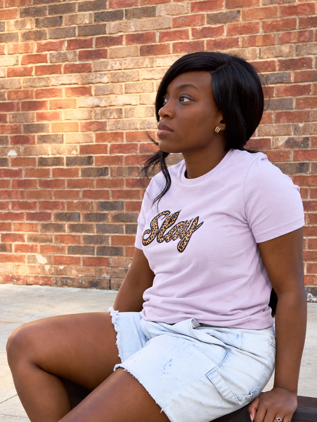 A woman sitting against a brick wall wearing a light pink t-shirt with 'Stay' written in leopard print script and light denim shorts.