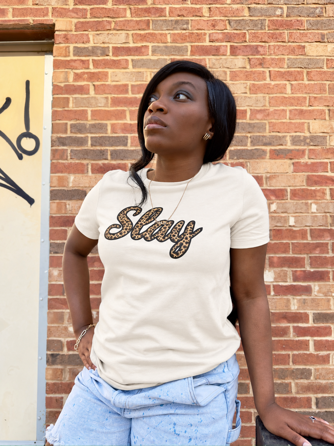 A woman wearing a cream-colored t-shirt with 'Slay' written in leopard-print cursive, paired with light blue jeans, standing against a red brick wall.