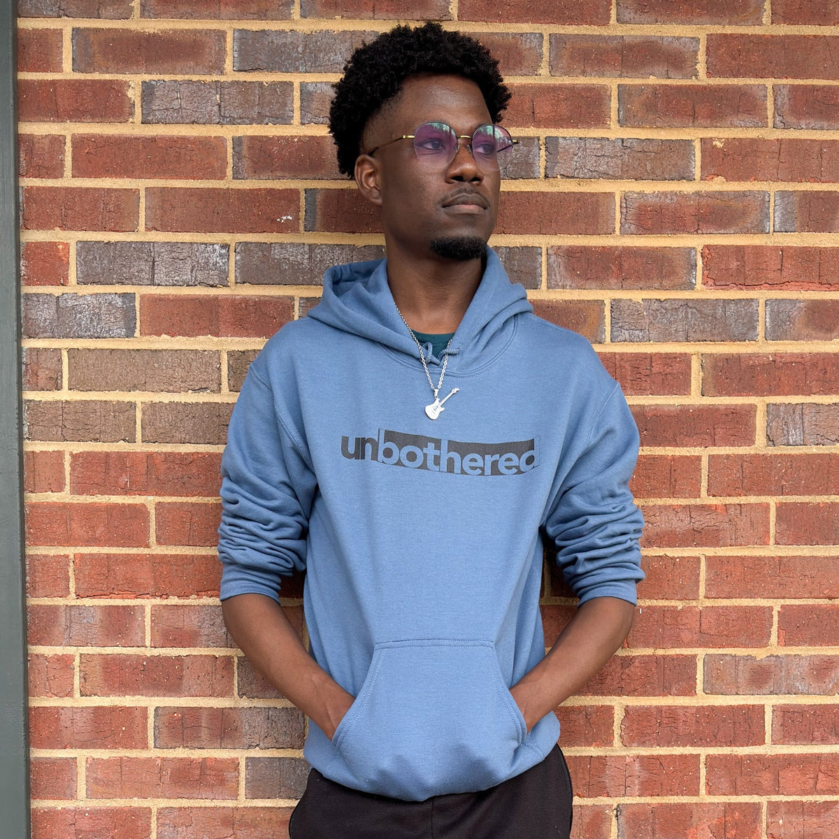 A man wearing a blue hoodie with 'unbothered' text against a brick wall