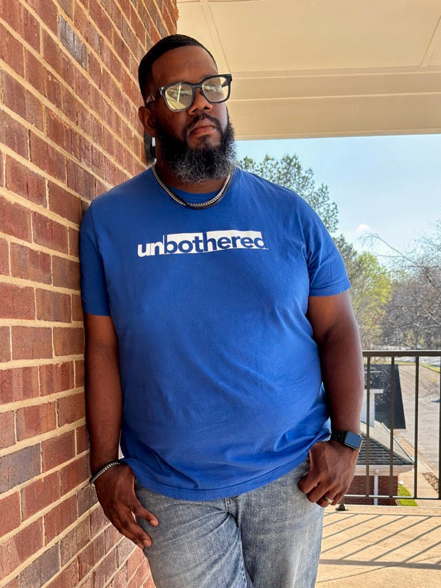 Man wearing a blue 'unbothered' t-shirt standing on a balcony.