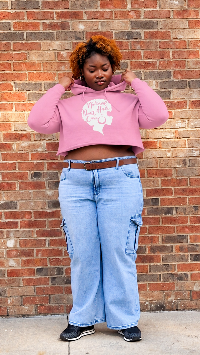 A woman stands in front of a red brick wall wearing a pink cropped hoodie with a white graphic of a silhouette of a woman with an afro, light blue wide-leg cargo jeans, a brown belt, and black sneakers.