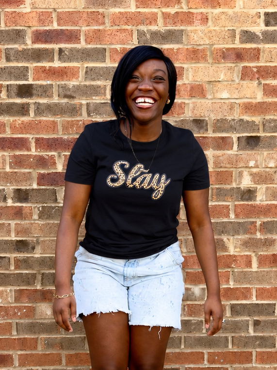 A woman stands in front of a brick wall wearing a black graphic t-shirt with the word 'Stay' in leopard script and light blue denim shorts.