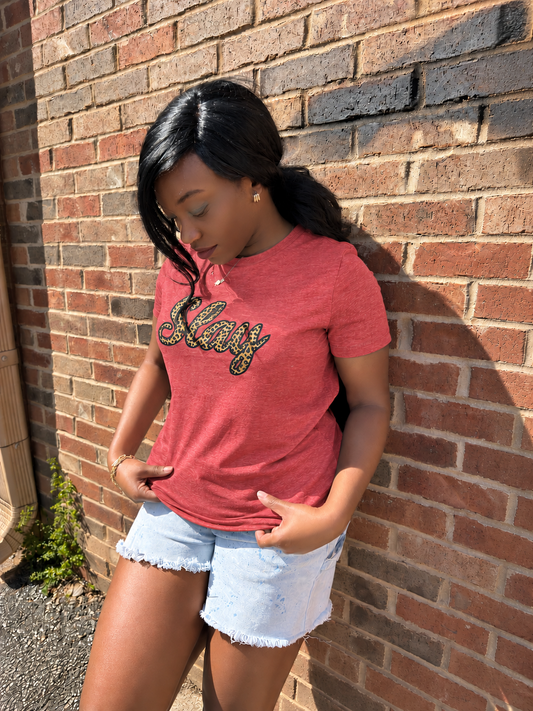 A woman wearing a heathered coral-red graphic t-shirt with 'Slay' in leopard and black lettering, paired with light-wash denim shorts, posing against a red brick wall.