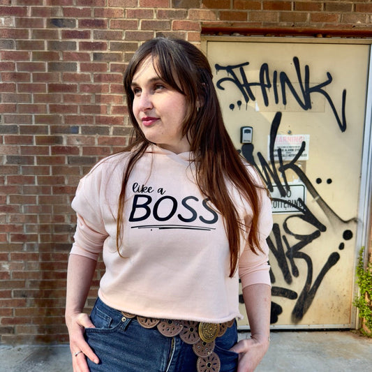 Woman wearing a 'like a BOSS' cropped hoodie standing in front of a brick wall with graffiti.