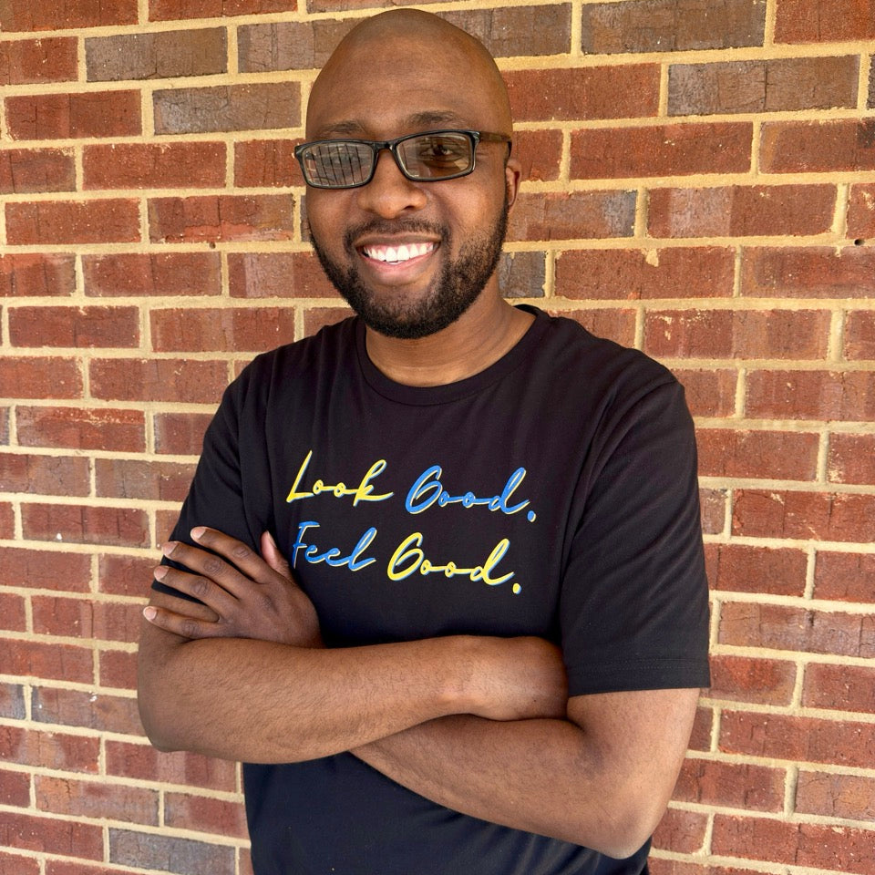 Man smiling wearing a black t-shirt with motivational text in a script font that says "Look Good. Feel Good." in yellow and blue lettering standing against a brick wall.