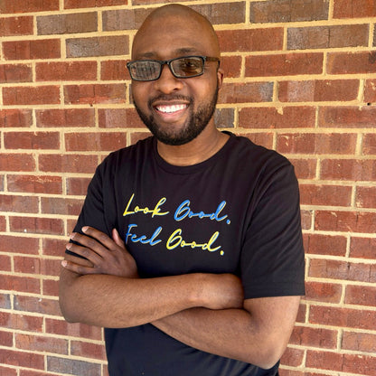 Man smiling wearing a black t-shirt with motivational text in a script font that says "Look Good. Feel Good." in yellow and blue lettering standing against a brick wall.