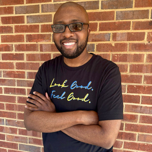 Man smiling wearing a black t-shirt with motivational text in a script font that says "Look Good. Feel Good." in yellow and blue lettering standing against a brick wall.