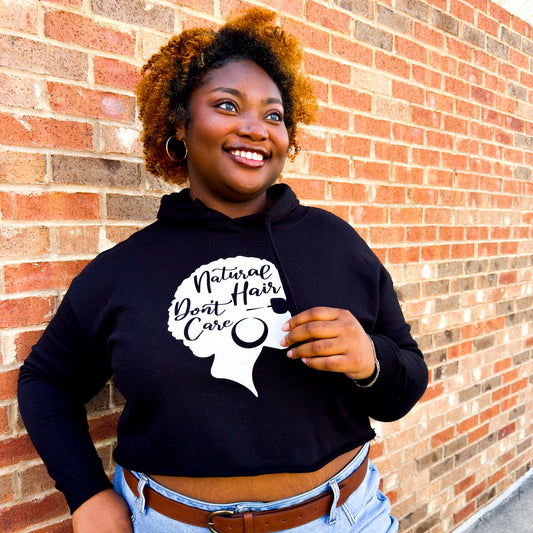 Woman smiling wearing a black cropped hoodie with a white graphic of a woman with a natural kinky afro and a hoop earring with the words "Natural Hair Don't Care" written inside.