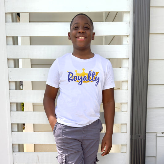 Young boy wearing a white t-shirt with 'Royalty' printed on it in a purple script font in front of a gold crown, standing in front of a white wooden fence.
