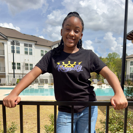 A smiling young girl, standing outside, wearing a black t-shirt that says "Royalty" in purple script in front of a gold crown.