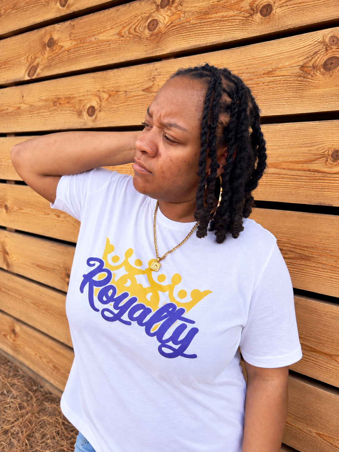 A woman with black braids leans against a wooden slat wall, wearing a white t-shirt with a yellow crown graphic and the word 'Royalty' in purple script.