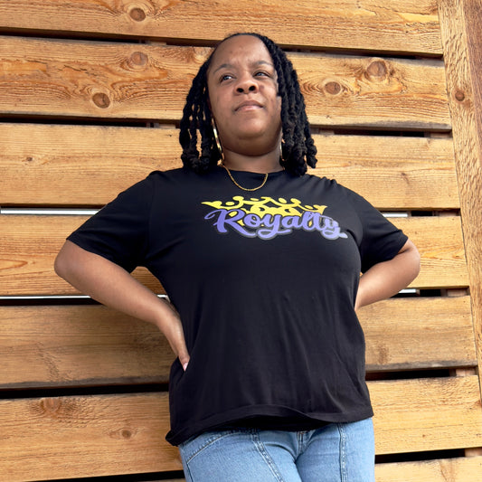Black woman wearing a black t-shirt with 'Royalty' printed on it in purple script font in front of a gold crown, standing against a wooden background
